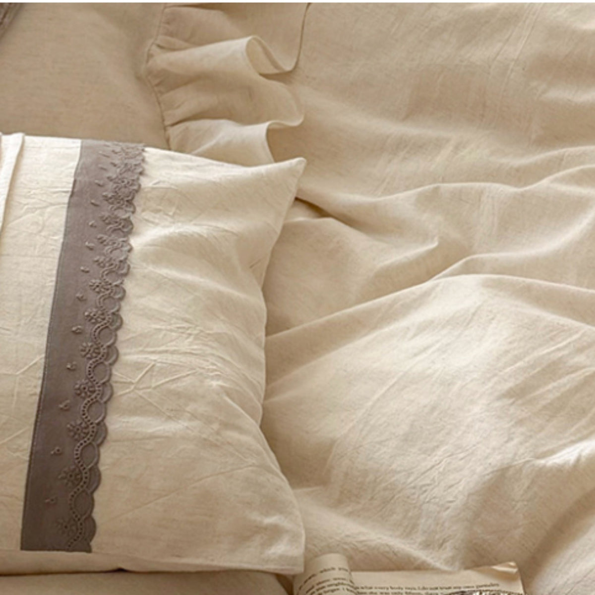 A close-up of a cream-colored bed linens set, featuring a textured pillowcase with a contrasting dusty gray scalloped lace trim, layered over matching rumpled sheets.