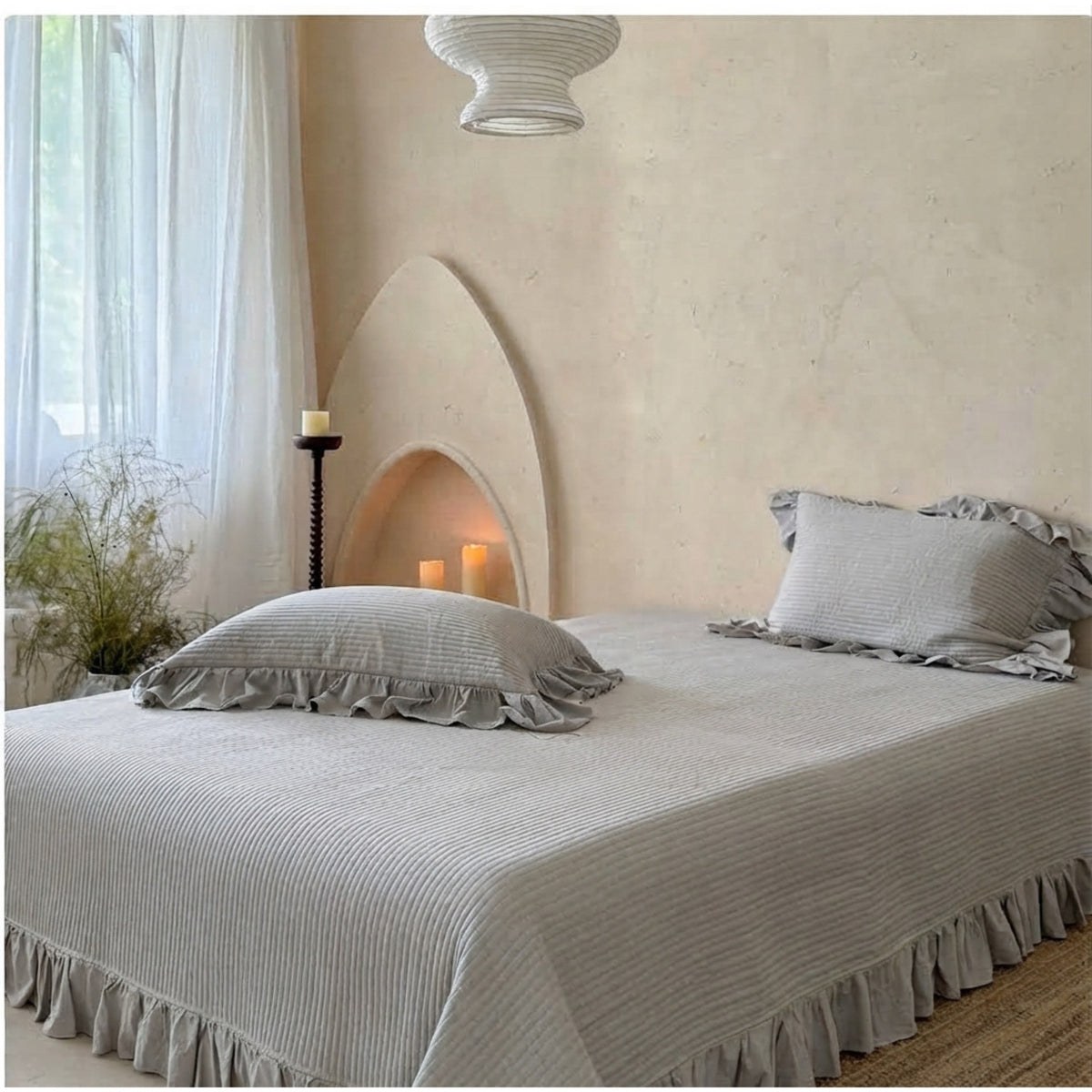 A bed dressed in a light gray ribbed bedspread and matching pillowcases with ruffled edges, styled in a softly lit bedroom with a textured beige wall, arched niche, candles, and sheer curtains.